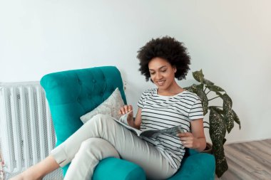 Güzel, genç, melez bir kadın koltukta oturuyor, dergi okuyor ve evde dinleniyor.