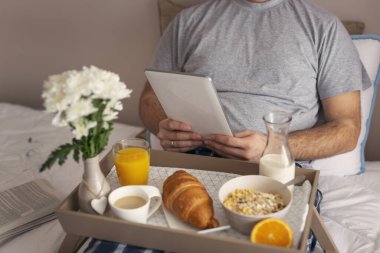 Pijama giyen bir adam, tablet bilgisayarda hazır haberler ve yatakta kahvaltı yapan bir adam.