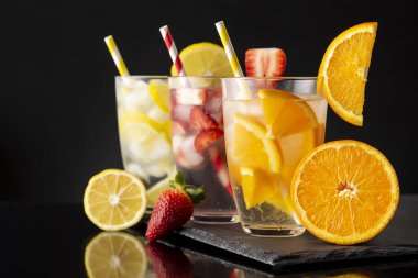 Siyah taş tepside taze limonlu, çilekli ve portakal dilimli soğuk su bardakları. Portakallı suya odaklan.