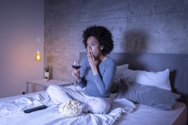 Güzel melez kadın yatakta oturuyor, şarap içiyor, patlamış mısır yiyor ve televizyon seyrediyor.