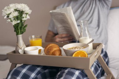 Pijama giyen, gazete okuyan ve yatakta kahvaltı yapan bir adam. Gevreğe odaklan.