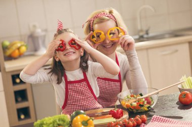 Güzel anne ve kız mutfakta salata yaparken, biber dilimlerini bardak gibi tutarken ve dikizlerken eğleniyorlar. Kızına odaklan.