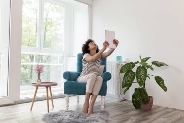 Güzel, genç, melez bir kadın koltukta oturuyor, tablet bilgisayarında video görüşmesi yapıyor, öpücükler yolluyor, evde boş zamanlarından zevk alan bir kadın.