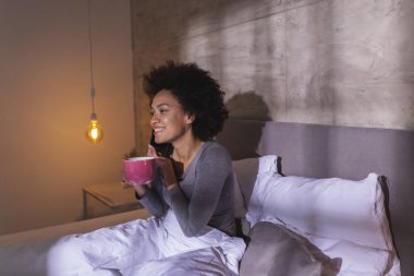 Neşeli, genç, melez bir kadın pijama giymiş, telefonla konuşuyor, kahve içiyor, sabahları evde dinleniyor.