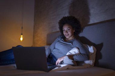 Genç ve güzel melez bir kadın gece yatağında dizüstü bilgisayarında video görüşmesi yapıyor.