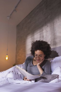 Güzel, melez, genç bir kadın pijamalarıyla yatakta uzanıyor, gazete okuyor ve sabahları evde boş vakitlerin tadını çıkarıyor.