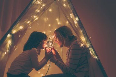 Anne ve kızı bir çadırda oturuyor, elinde bir kavanozla yılbaşı ışıklarıyla hikayeler anlatıyor. Annesine odaklan.