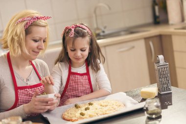 Anne ve kızı mutfakta pizza yapıyor, küçük kız pizzanın üstüne zeytin koyuyor, pişirmeye hazır. Kızına odaklan.