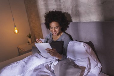 Sabahları pijama giyen, tablet bilgisayar kullanan, gülen, el sallayan ve evde dinlenen güzel, melez bir kadın.