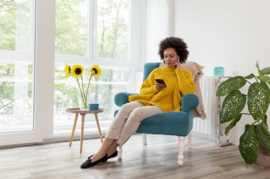 Güzel melez bir kadın koltukta oturuyor, evde dinleniyor, akıllı telefondan mesaj yazıyor.