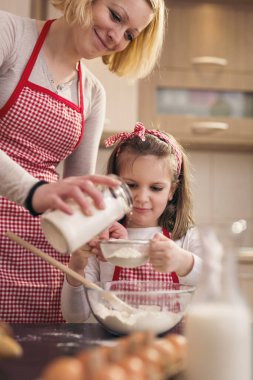 Anne ve kızı hamur pişirirken, anne eleğe biraz daha un eklerken, kız da elek ile yoğurma kabına un ekiyor.