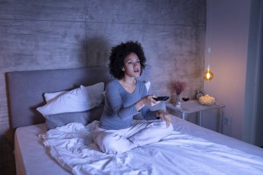 Yatakta pijama giyen, patlamış mısır yiyen ve uzaktan kumandayla TV 'de kanal değiştiren güzel melez bir kadın.