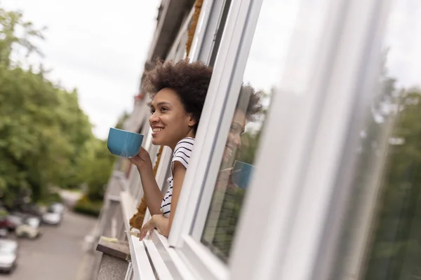 Beautiful young mixed race woman standing by the window and looking out on the street, relaxing at home and drinking coffee, enjoying her leisure time