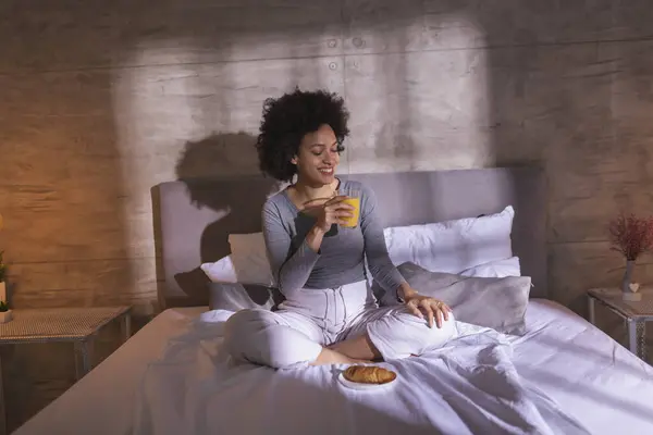 Beautiful young mixed race woman wearing pajamas sitting on bed, having breakfast and relaxing at home in the morning
