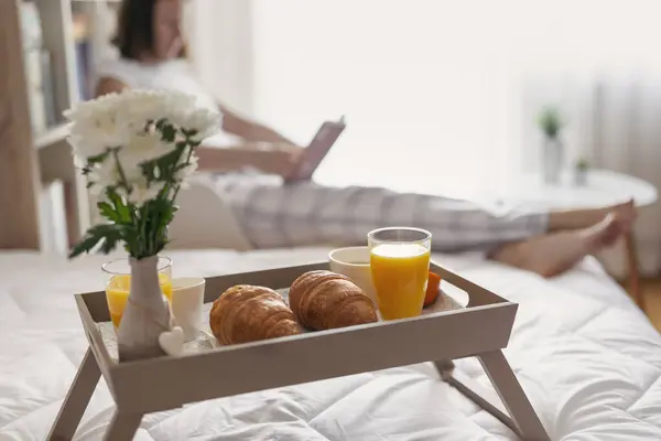 穿着睡衣的女人坐在床边的椅子上看书早餐托盘在前景 有选择地关注右边的羊角面包 — 图库照片