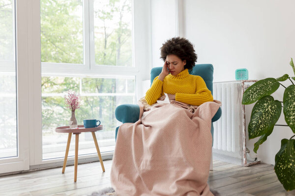 Portrait of sick, tired woman sitting in an armchair covered with blanket, having migraine, holding head in hands