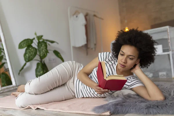 Beautiful young mixed race woman lying on the bedroom floor, reading a book and relaxing at home