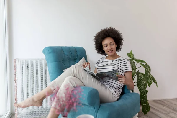 Güzel, genç, melez bir kadın pencerenin kenarında oturmuş dergi okuyor ve evde eğleniyor.