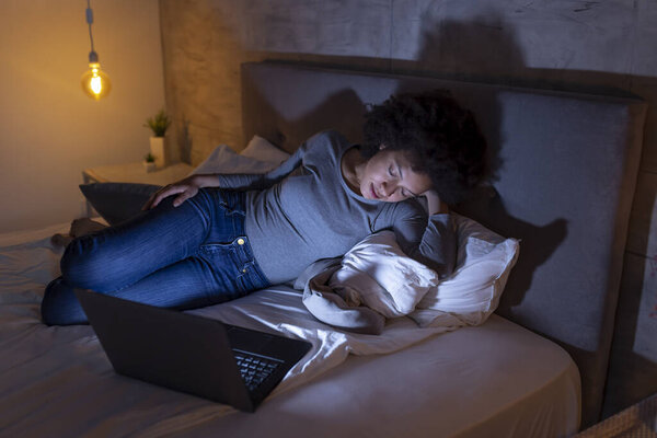 Красивая молодая смешанная расовая женщина, лежащая в постели, заснула во время просмотра фильма на ноутбуке