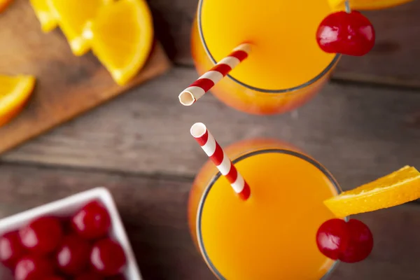 Table top shot of two cold tequila sunrise cocktails with tequila, pomegranate juice and orange juice decorated with slices of orange and maraschino cherries. Focus on the tips of the straws