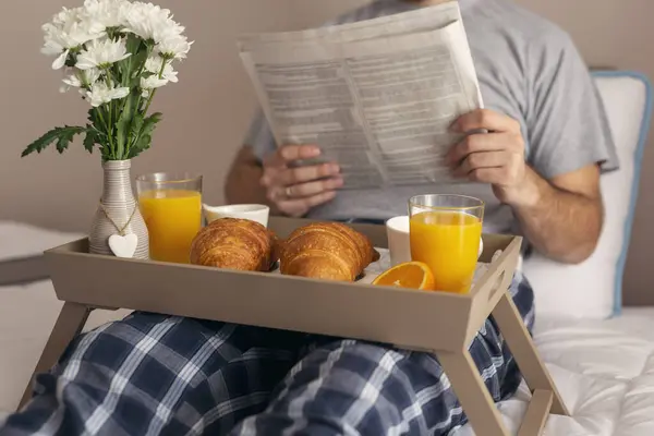 Man wearing pajamas, reading newspapers and having breakfast in bed. Selective focus on the croissants and the vase