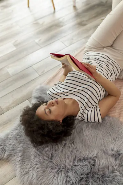 一个漂亮的男女混血的年轻女子躺在卧室的地板上 一边看书 一边在家里放松 她的高角镜 — 图库照片