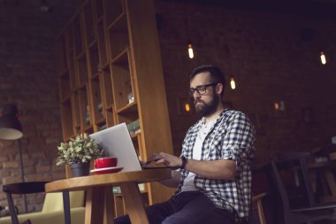 Yakışıklı hippi bir kafede koltukta oturuyor, sabah kahvesini içiyor ve serbest çalışmak için dizüstü bilgisayar kullanıyor.