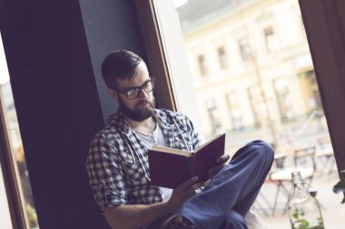 Young man sitting next to a window in a cafe, reading a book and enjoying his leisure time clipart