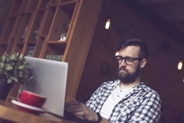 Yakışıklı hippi adam kafede oturuyor, sabah kahvesini içiyor ve bilgisayarını serbest çalışmak için kullanıyor.