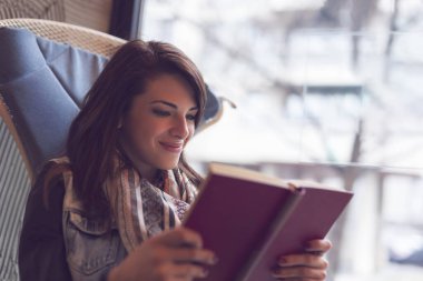 Pencerenin yanındaki kafede oturan, gülümseyen ve kitap okuyan güzel genç bir kadın.