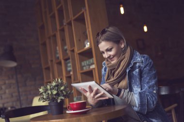 Güzel, genç bir sarışın kafeteryada dalgın bir şekilde oturuyor, sabah kahvesini içiyor, elinde tablet bilgisayarla internette sörf yapıyor.