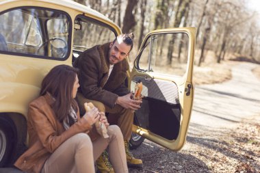 Genç bir çift araba yolculuklarına ara verip, yol kenarında oturup sandviç yiyorlar. Adama odaklan.