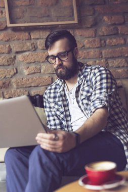 Yakışıklı hippi, bir kafede kanepede oturuyor, sabah kahvesini içiyor ve serbest çalışmak için dizüstü bilgisayar kullanıyor.