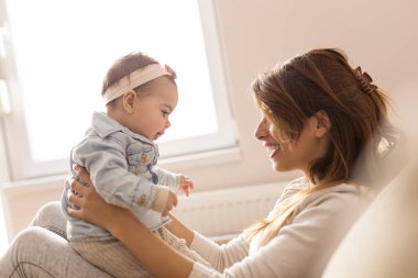 Genç anne bebeğini kollarında tutuyor, oturma odasındaki kanepede oturuyor, pencerenin yanında gülüyor ve oynuyor.