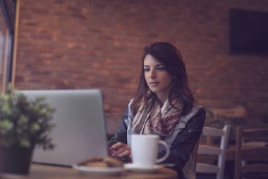 Genç bir kadın kafede oturuyor, bir fincan kahve içiyor ve dizüstü bilgisayar kullanıyor.