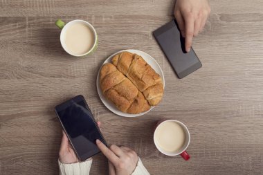 Kadın ve erkeğin elinde akıllı telefonlar, internette gezinirken kahve içerken ve kahvaltı yaparken görüntüsü.