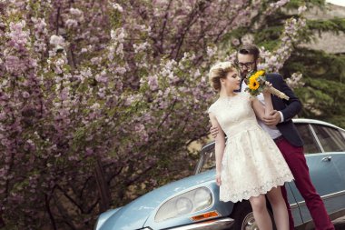 Yeni evli çift eski bir arabanın yanında duruyor. Balayına çıkıp kucaklaşıyorlar.