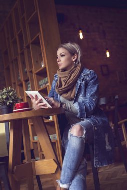 Güzel genç bir sarışın kafeteryada dalgın bir şekilde oturuyor, sabah kahvesini içiyor, elinde bir tablet bilgisayar tutuyor ve internete yüklüyor.