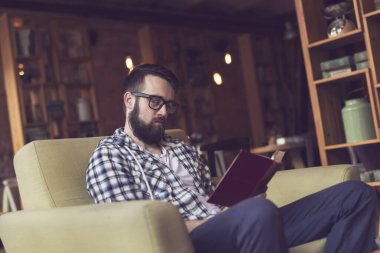 Genç adam bir kafede rahat bir koltukta oturuyor, kitap okuyor ve boş zamanlarının tadını çıkarıyor.