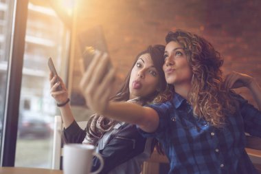 İki arkadaş bir kafede kahve içiyor, akıllı bir telefonla selfie çekiyor ve komik suratlar yapıyor. Soldaki kıza odaklan.