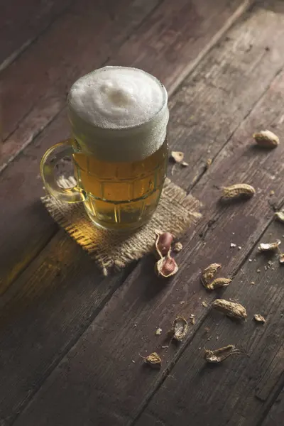Mug of light beer with froth placed on a burlap cover and some peanuts on a rustic wooden pub table. Focus on the froth