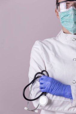 Üniformalı doktor, koruyucu eldiven takıyor, yüz koruma maskesi ve gözlük takıyor ve steteskop tutuyor.