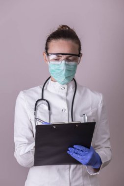 Üniformalı doktor, tıbbi yüz koruma maskesi, koruyucu eldiven ve gözlük takıyor, yazı tahtası tutuyor ve not alıyor; covid-19 salgınına karşı savaşan tıbbi personel
