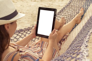 Tatilde plajda bir hamakta uzanmış, elinde boş ekranlı bir tablet bilgisayar tutan bir kadın.