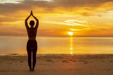 Sahilde yoga yapan aktif genç bir kadın, elinde ağaç pozu ya da Vrikshasana, yoga pozisyonunu dengeliyor ve suda güzel bir gün doğumunun tadını çıkarıyor.