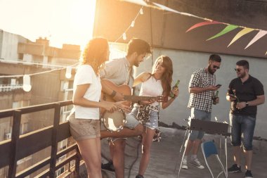 Arkadaşlar çatıdaki partide eğleniyor, gitar çalıyor ve şarkı söylüyorlar. Gitar çalan adama odaklan.