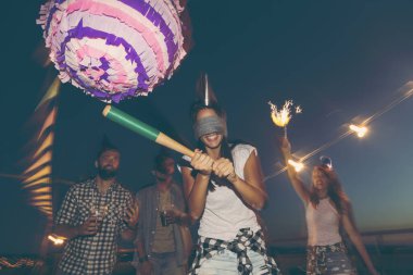 Birthday girl hitting the pinata with baseball bat while her friends are cheering and laughing. Young people having fun at a rooftop birthday party clipart