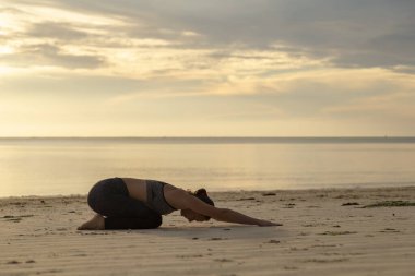 Genç bir kadın sabahın erken saatlerinde plajda antrenman yapıyor, yoga yapıyor ve esniyor, Balasana ya da çocuk pozu veriyor.