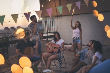 Genç arkadaşlar yazlık çatı partisinde eğleniyor, gitar çalıyor, şarkı söylüyor, dans ediyor ve dinleniyorlar. Ortadakilere odaklan.
