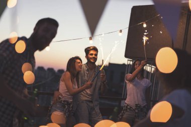 Bir grup genç arkadaş çatıdaki bir partide eğleniyor, şarkı söylüyor, dans ediyor ve maytap sallıyorlar.